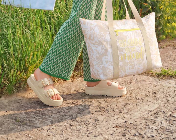 Close-up of beige platform slides worn with green patterned pants and a tote bag beside the legs.
