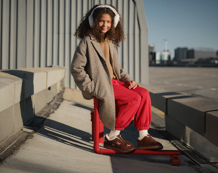 Person sitting on a red metal rail in an outdoor urban setting, wearing a checked oversized coat, red trousers, earmuffs, and brown slip-on shoes. Modern buildings and open sky are visible in the background.