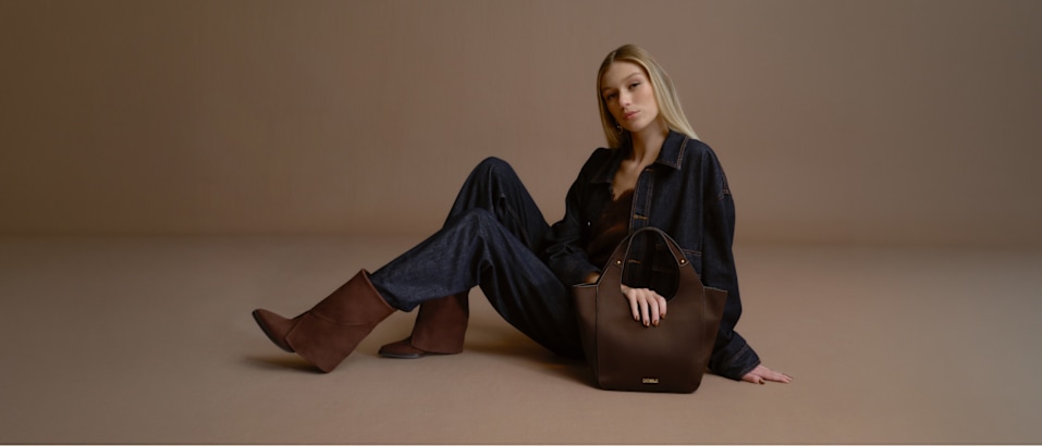 A studio image featuring a seated model on a neutral beige background, styled in a dark denim outfit paired with brown ankle boots. Beside the model is a structured dark brown handbag with short handles. The coordinated fashion look combines rich brown tones and denim textures, creating a clean autumn-inspired presentation with a minimalist backdrop.