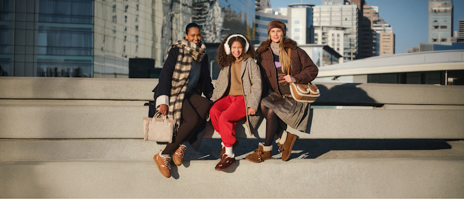 Three people sitting on concrete steps in an urban setting, wearing seasonal outfits and casual shoes. Modern city buildings are visible in the background.