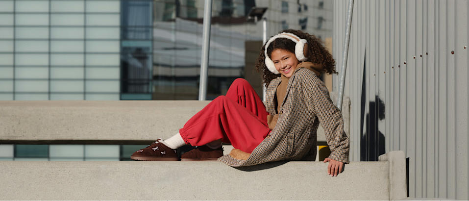 Person standing outdoors on a concrete ledge in front of a modern building, wearing white earmuffs, a checked oversized coat, red trousers, and brown slip-on shoes.