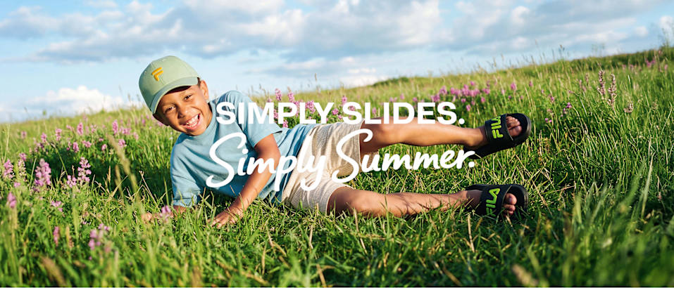 Child lying in grass wearing black slide sandals with green logo detail. Text reads “SIMPLY SLIDES. Simply Summer.”