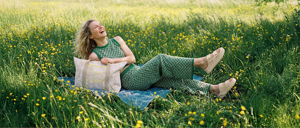 Person lying on a blanket in a field wearing beige clog-style shoes and holding a patterned bag.