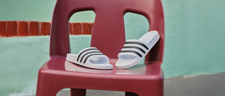A pair of white Adidas slides with black stripes is displayed on a red plastic chair.