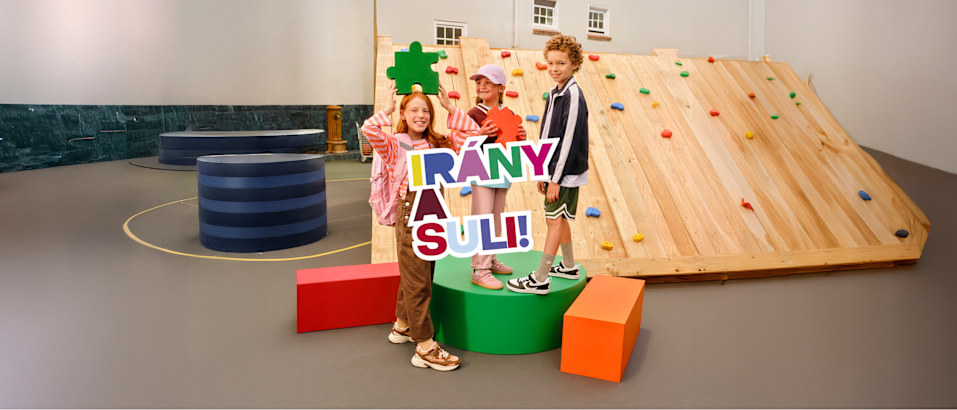 Children posing with colorful school-themed letters in front of a climbing wall. Letters say "Irany a Suli".