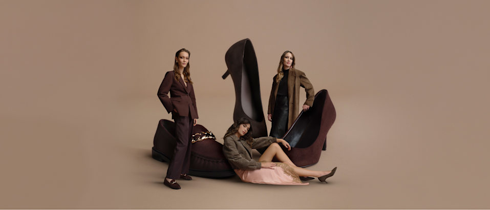 Fashion campaign image on a beige background featuring oversized chocolate brown loafers and high heels. Three models are posed around the shoes, wearing coordinated outfits in brown, black, and neutral tones. The footwear and styling create a sophisticated autumn fashion scene with a focus on rich brown shades.