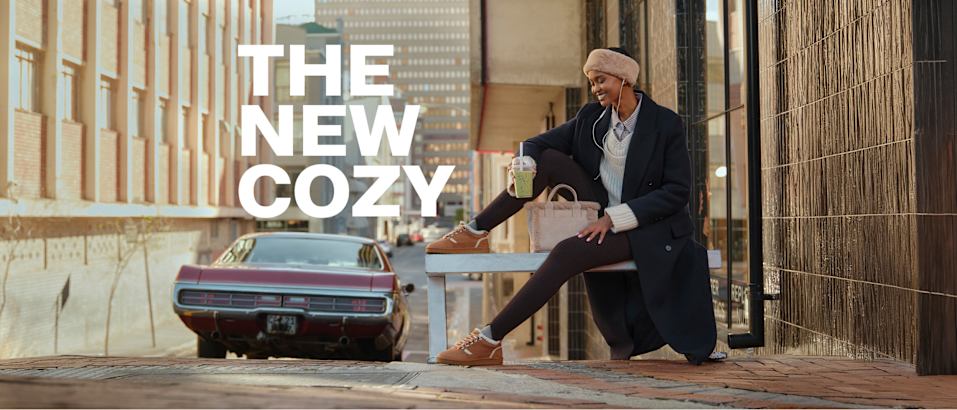 Person sitting on a white bench in a city street setting, wearing a dark overcoat, light knit hat, sweater, skirt, tights, and brown lace-up shoes. Large white text on the left reads: 'THE NEW COZY'. A vintage car is parked in the background.
